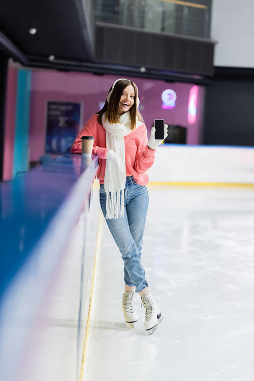 full length of happy woman in ear muffs and gloves holding smartphone with blank screen on ice rink