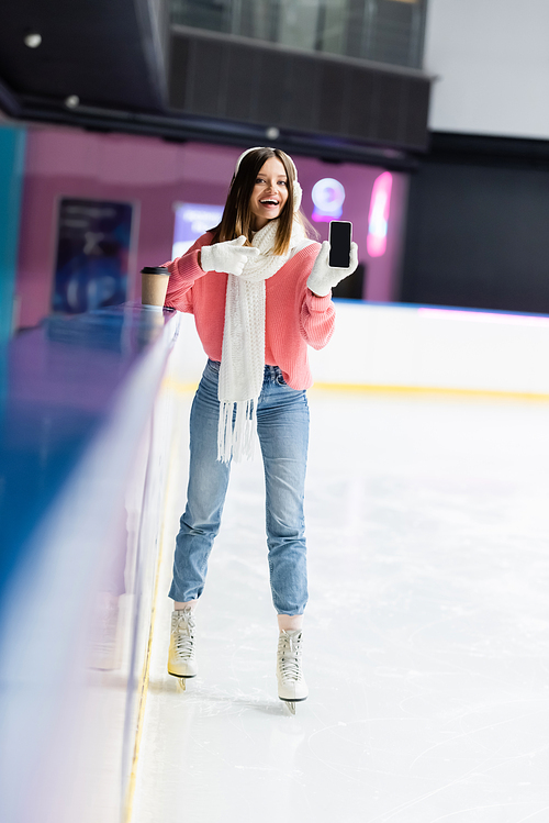 happy woman in white ear muffs and pink sweater pointing with finger at cellphone with blank screen on ice rink