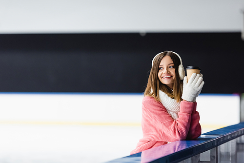 happy woman in knitted sweater, ear muffs and winter outfit holding paper cup on ice rink