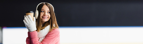 cheerful young woman in ear muffs and scarf holding paper cup on ice rink, banner