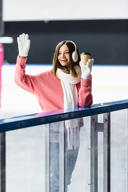 excited woman in ear muffs and pink sweater holding paper cup and waving hand on ice rink