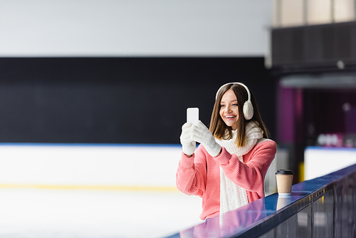 happy woman in ear muffs and gloves taking photo on ice rink