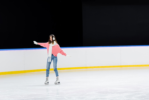 full length of excited young woman in ear muffs and scarf skating with outstretched hands on ice rink