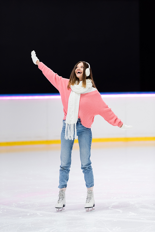 full length of happy woman in ear muffs and winter outfit skating on ice rink