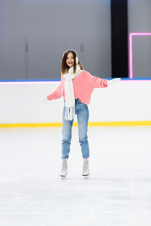 positive young woman in ear muffs and scarf skating with outstretched hands on ice rink