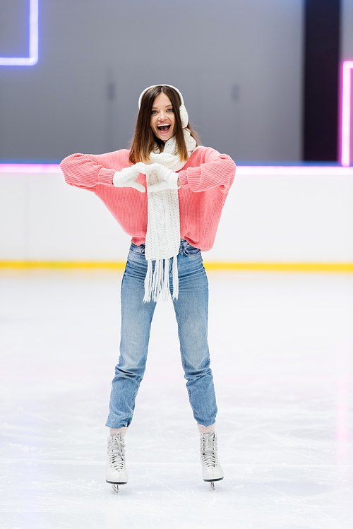 full length of cheerful woman in white scarf and ear muffs showing heart with hands while skating on ice rink