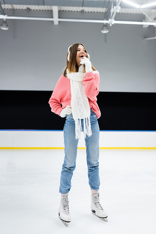 full length of cheerful woman in white ear muffs and knitted scarf skating on ice rink