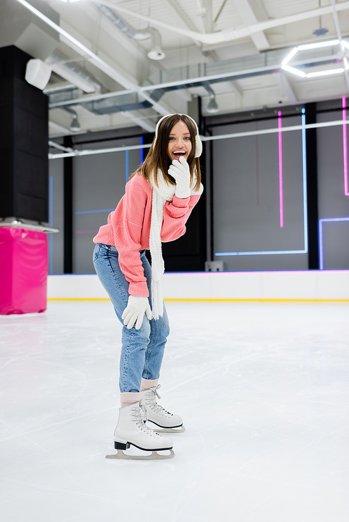 full length of cheerful woman in white ear muffs and knitted scarf skating while laughing on ice rink