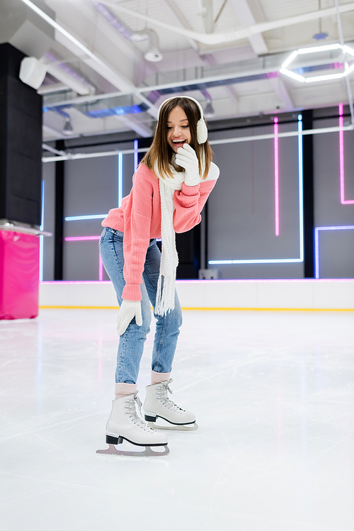 full length of happy young woman in ear muffs and scarf skating while laughing on ice rink