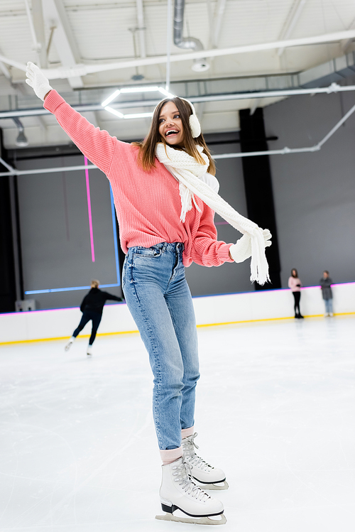 full length of excited woman in white ear muffs holding scarf while skating with outstretched hand on ice rink
