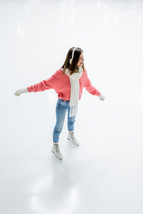 high angle view of pleased young woman in ear muffs and scarf skating on ice rink