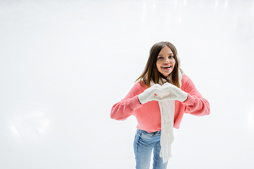 happy young woman in white ear muffs and knitted scarf showing heart sign on ice rink