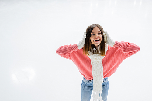high angle view of excited young woman in scarf adjusting white ear muffs on ice rink