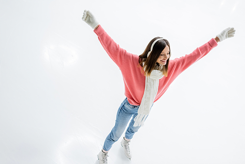 high angle view of of excited young woman in ear muffs and jeans skating with outstretched hands on ice rink