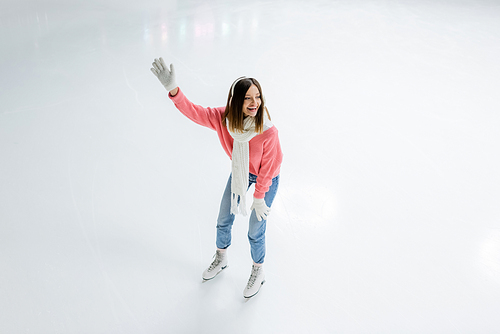 high angle view of amazed young woman in white ear muffs and knitted sweater skating with outstretched hand on ice rink