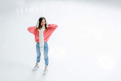 high angle view of amazed young woman holding white ear muffs and standing on ice rink