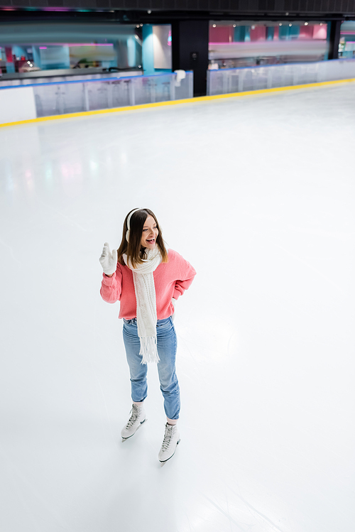 full length of happy woman posing with hand on hip and waving hand on ice rink