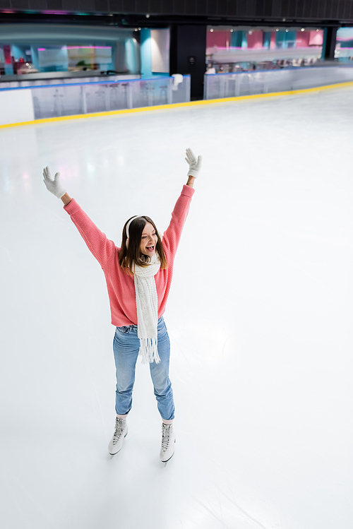 high angle view of amazed young woman in white ear muffs skating with raised hands on ice rink