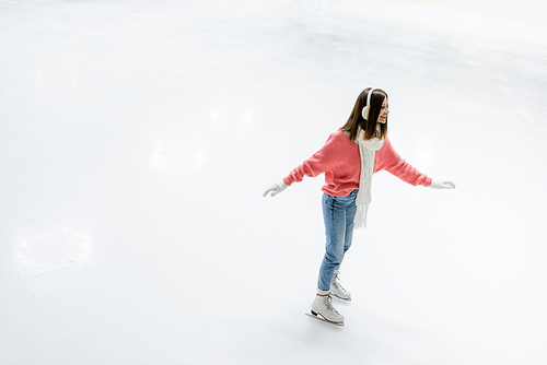 high angle view of cheerful young woman in ear muffs and scarf skating on ice rink