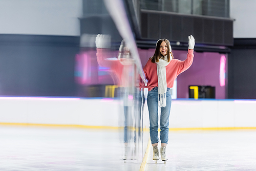 pleased woman in white ear muffs and pink sweater skating and waving hand on ice rink