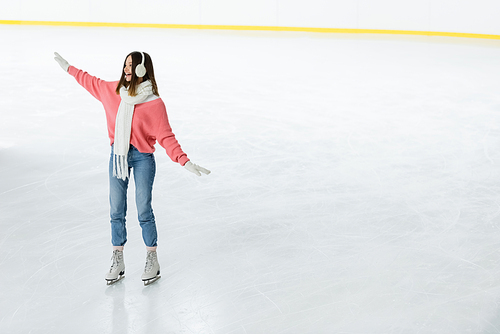full length of amazed young woman in ear muffs and scarf skating with outstretched hands on ice rink