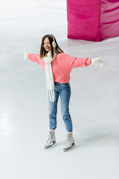 full length of joyful young woman in ear muffs and knitted scarf skating with outstretched hands on ice rink