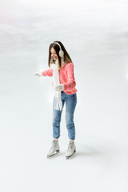 full length of positive young woman skating with outstretched hands on frozen ice rink