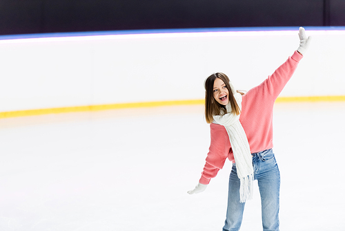 amazed young woman in white ear muffs and knitted scarf skating with outstretched hands on ice rink
