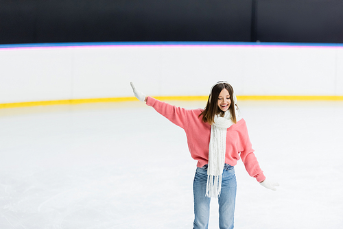 smiling woman in white ear muffs, scarf and sweater skating with outstretched hand on ice rink