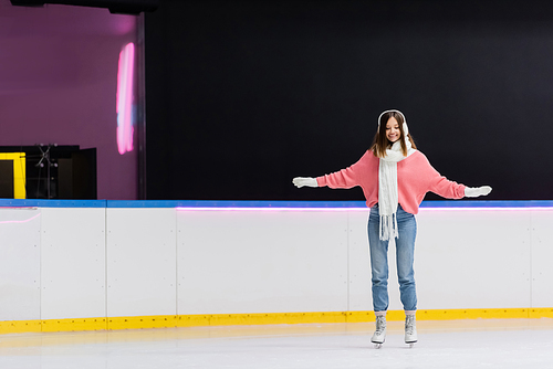 full length of cheerful woman in white ear muffs and knitted scarf skating with outstretched hands on ice rink