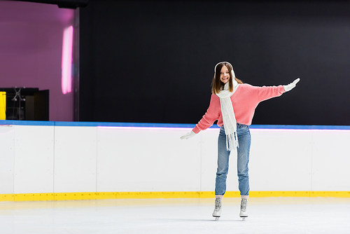 full length of smiling woman in ear muffs and winter outfit skating on ice rink