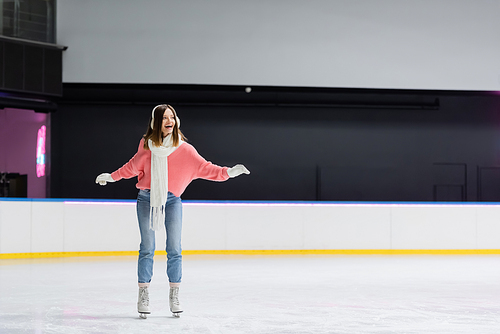 full length of astonished woman in white scarf, sweater and ear muffs skating on ice rink