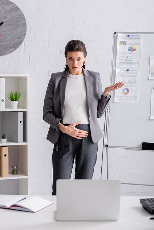 young pregnant businesswoman gesturing near flip chart with chats and graphs during video call on laptop
