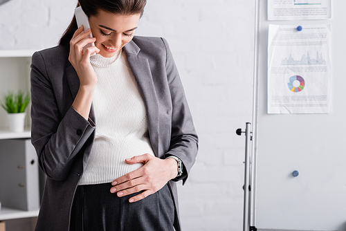 joyful pregnant businesswoman talking on smartphone near flip chart in office