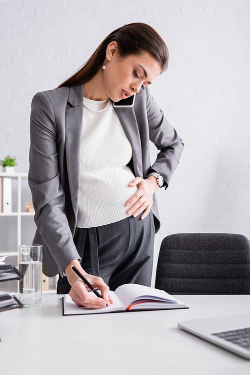 pregnant businesswoman writing in notebook and talking on cellphone