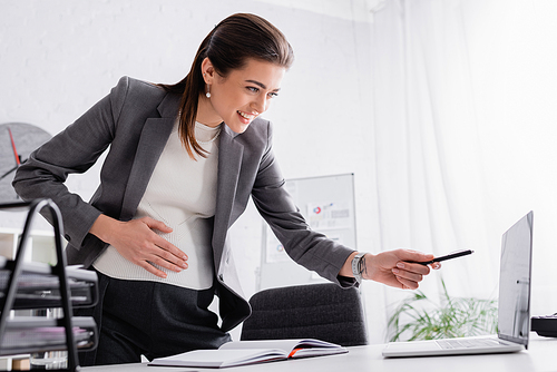 happy pregnant businesswoman pointing with pen at laptop on desk