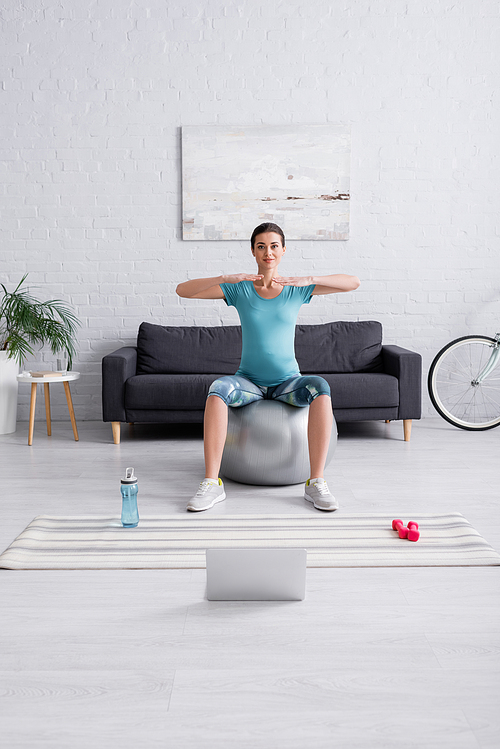 happy pregnant woman in sportswear exercising on fitness ball near laptop in living room