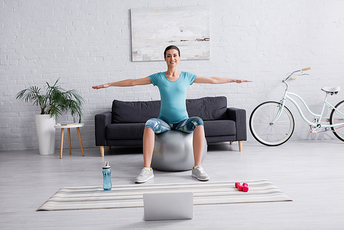cheerful pregnant woman in sportswear exercising on fitness ball near laptop in living room