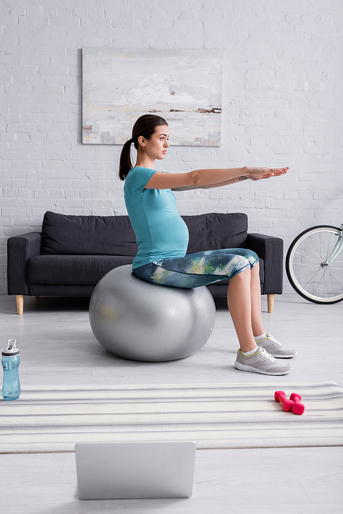 pregnant woman in sportswear exercising on fitness ball near laptop in living room