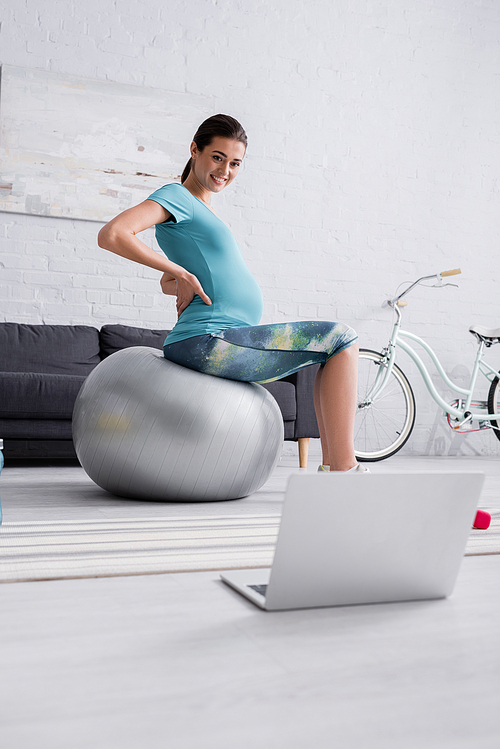 positive pregnant woman in sportswear exercising on fitness ball and looking at laptop in living room