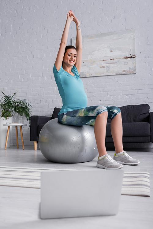 positive pregnant woman with raised hands exercising on fitness ball and looking at laptop in living room