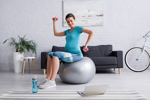 cheerful pregnant woman exercising on fitness ball with dumbbells near laptop