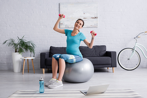 happy pregnant woman exercising on fitness ball with dumbbells near laptop