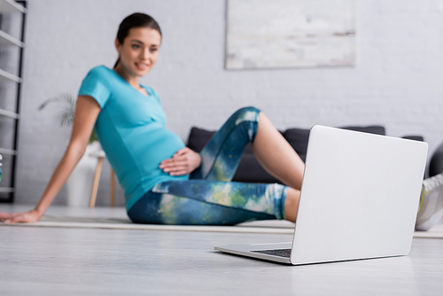 cheerful pregnant woman in sportswear sitting on fitness mat near laptop in living room