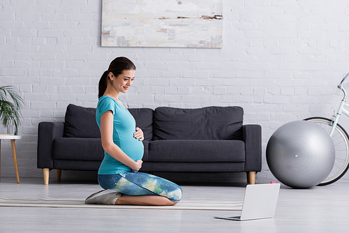 happy pregnant woman in sportswear working out near laptop on fitness mat