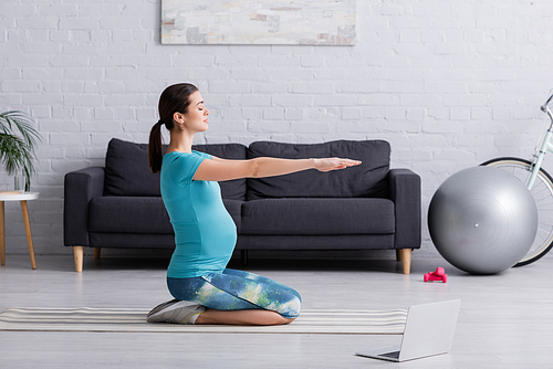 side view of pregnant woman in sportswear working out near laptop on fitness mat