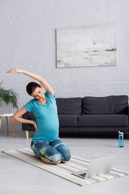 cheerful pregnant woman in sportswear stretching with hand on hip while looking at laptop
