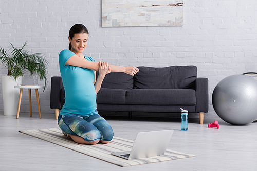 happy pregnant woman in sportswear stretching while looking at laptop