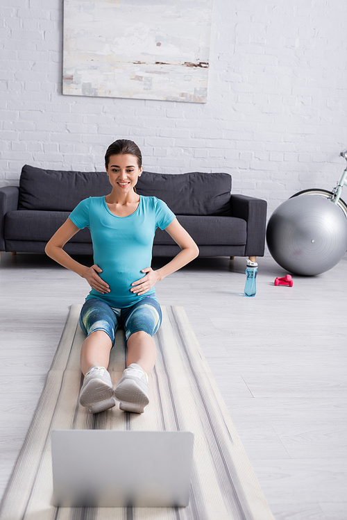 smiling pregnant woman in sportswear exercising near laptop in living room