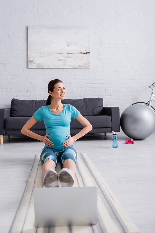 happy pregnant woman in sportswear exercising near laptop on fitness mat